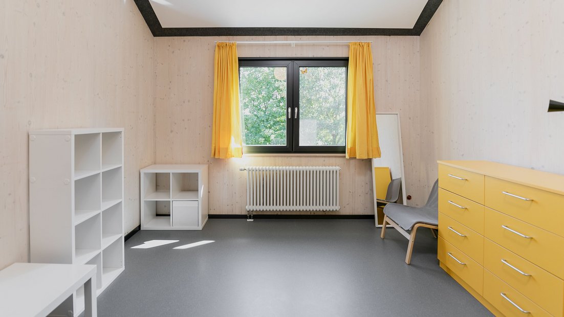 Chambre dans le Rosenhaus à l’étage avec commode jaune, étagères blanches, table, miroir et fauteuil gris